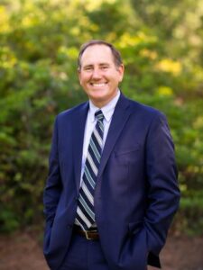 Smiling man in a navy suit and striped tie stands outdoors with greenery in the background.