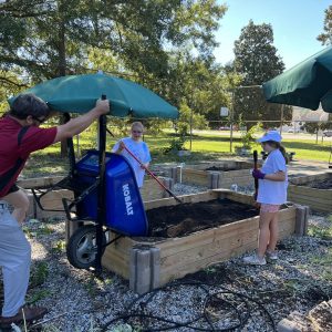 A group of people are working together to build garden boxes for the community. There is an adult using a wheelbarrow to pour soil into the garden box, and two girls are spreading the soil around to make sure it's evenly distributed.