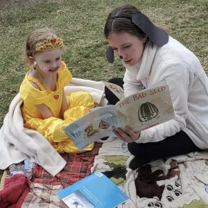 Imagine a young girl dressed in a beautiful princess costume, sitting on a blanket next to her mom. Her mom is dressed up as a dog and is also reading a book.