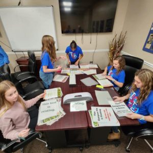 A group of young girls wearing all blue shirts are actively participating in creating a sign as part of the Ready Readers program. In the background, there is a television with wires hanging down, and to the left, there is a smart board. There are all black chairs and four desks arranged together.