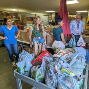 A group of kind volunteers are working together to carry food and provide assistance to the homeless. They are currently located in a warehouse filled with an abundance of food.